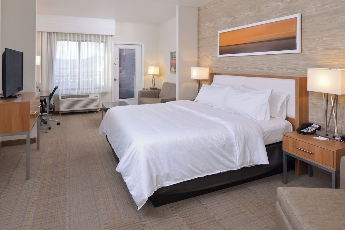A hotel room with a large bed, a desk with a computer, a chair, a TV, and a wall-mounted painting.