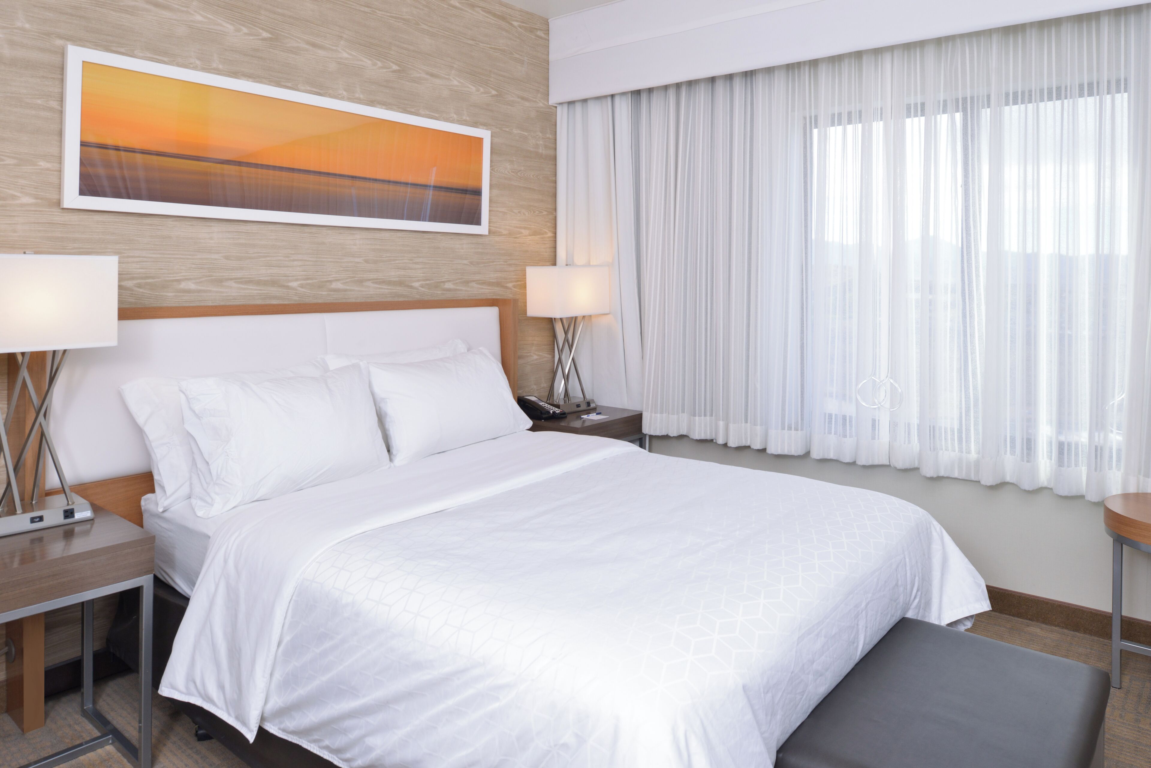 A hotel room with a large bed, two bedside tables with lamps, a nightstand, a window with sheer curtains, and a painting on the wall.