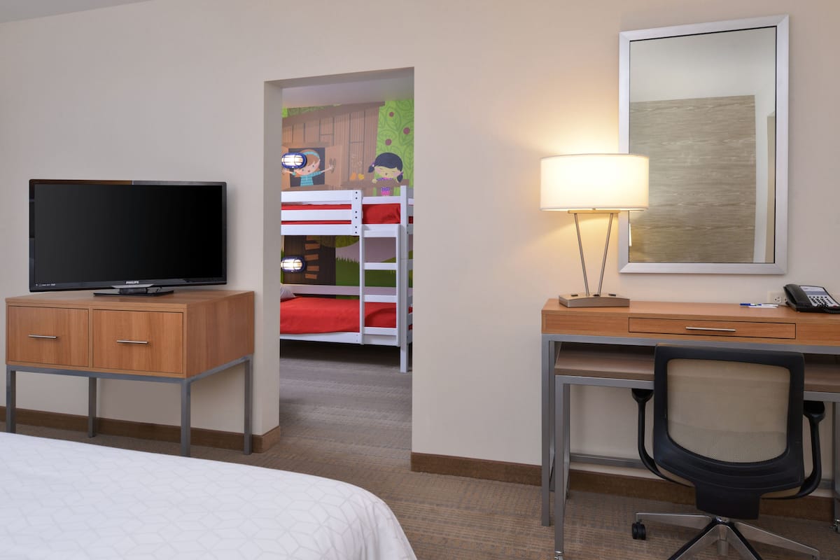 A hotel room with a bed, a desk with a lamp, a television on a wooden cabinet, and a bunk bed in the background.