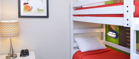 A bunk bed with a red bedspread, a framed picture on the wall, a lamp, and a game controller on a nightstand.