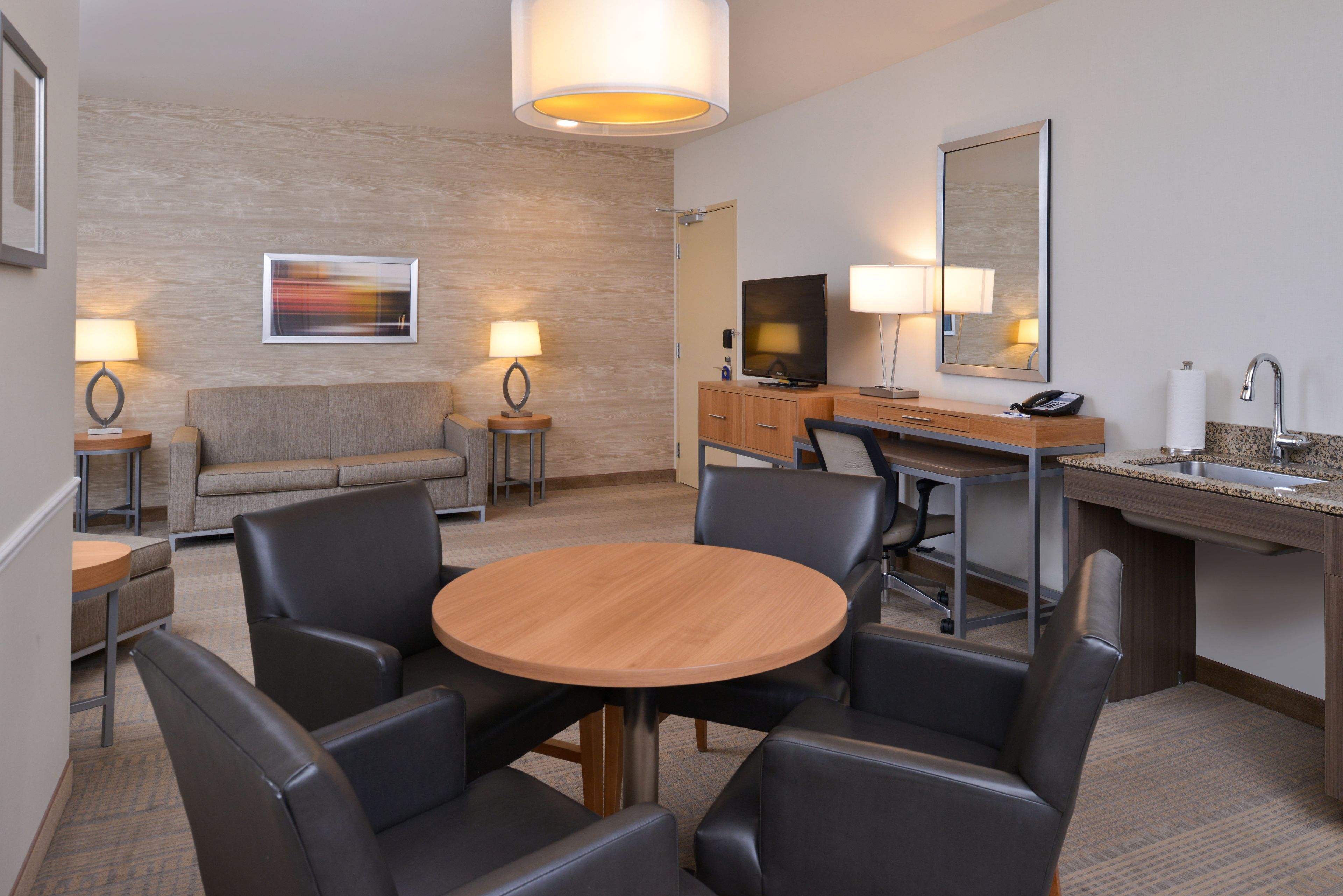 A hotel room with a round wooden table, four chairs, a sofa, a television, a desk with a mirror, and two lamps.