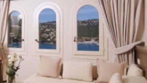 A room with a sofa, a table with a vase, and three arched windows overlooking a coastal view. - Hotel Villa Italia (Andraitx)
