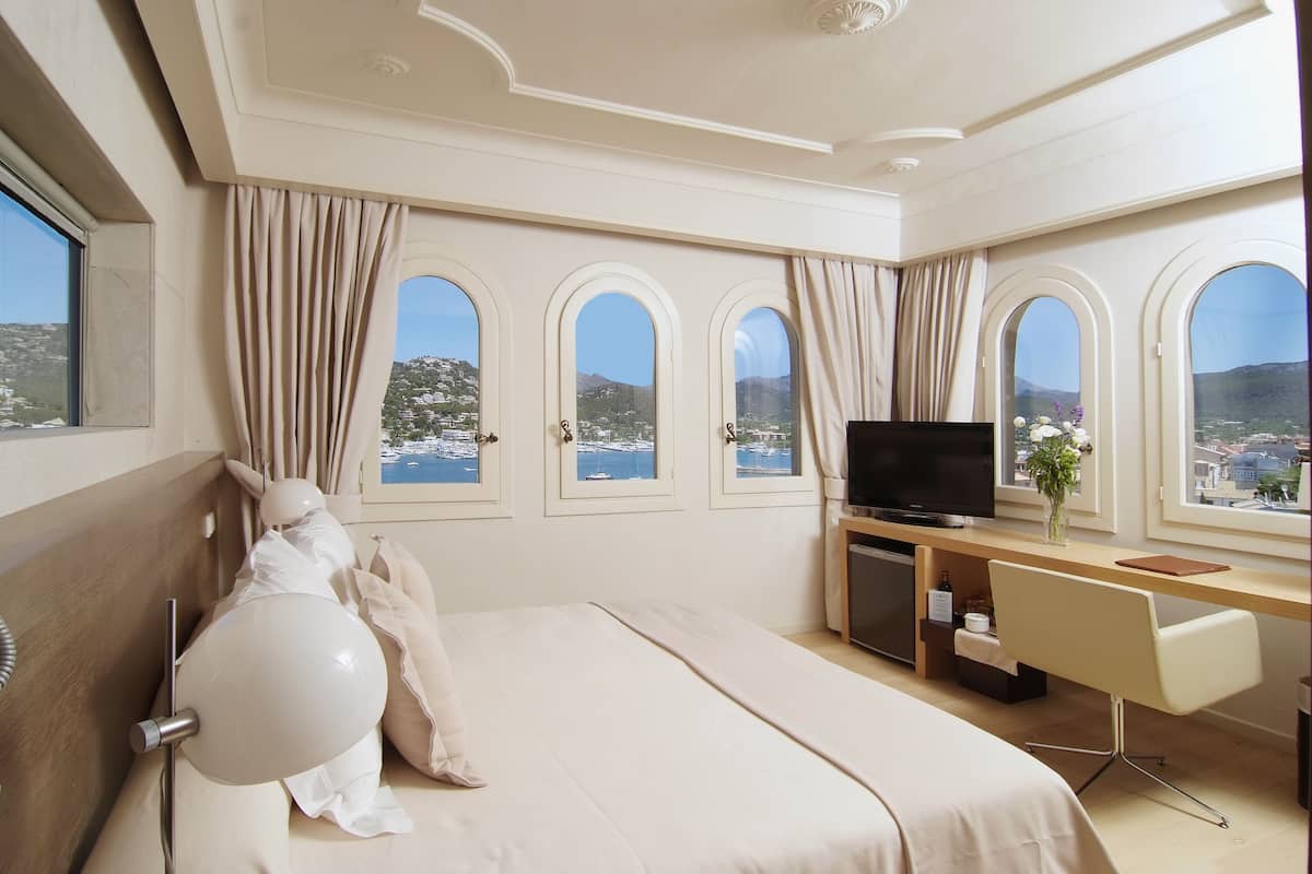 A hotel room with a large bed, a desk with a TV, and three arched windows with curtains.
