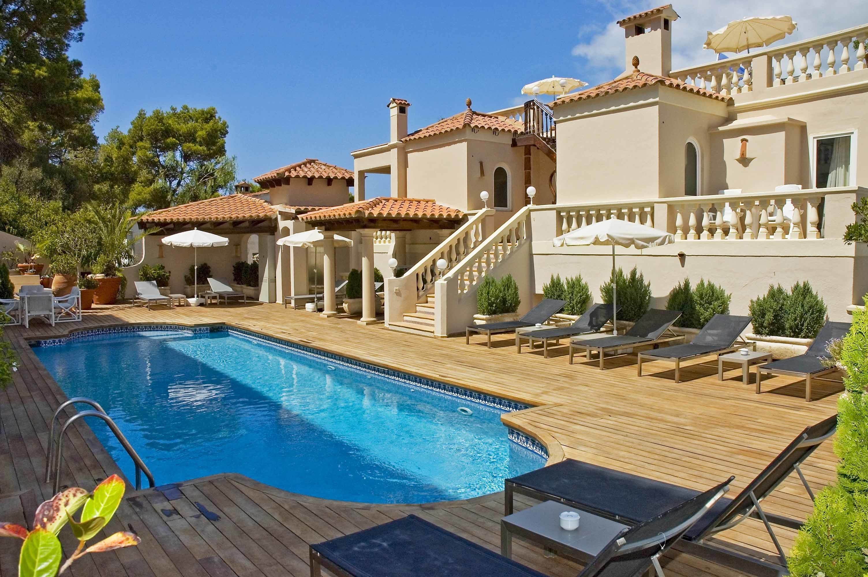 A swimming pool with lounge chairs and umbrellas, surrounded by a wooden deck and a building with a tiled roof.