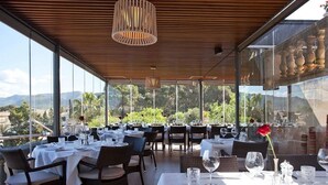 An outdoor dining area with tables set for a meal, large windows offering a view of the landscape, and hanging pendant lights. - Hotel Villa Italia (Andraitx)