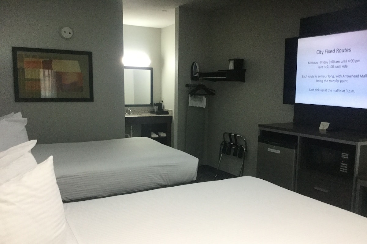 A hotel room with two beds, a TV displaying city fixed routes, a mirror, a sink, and a small cabinet.