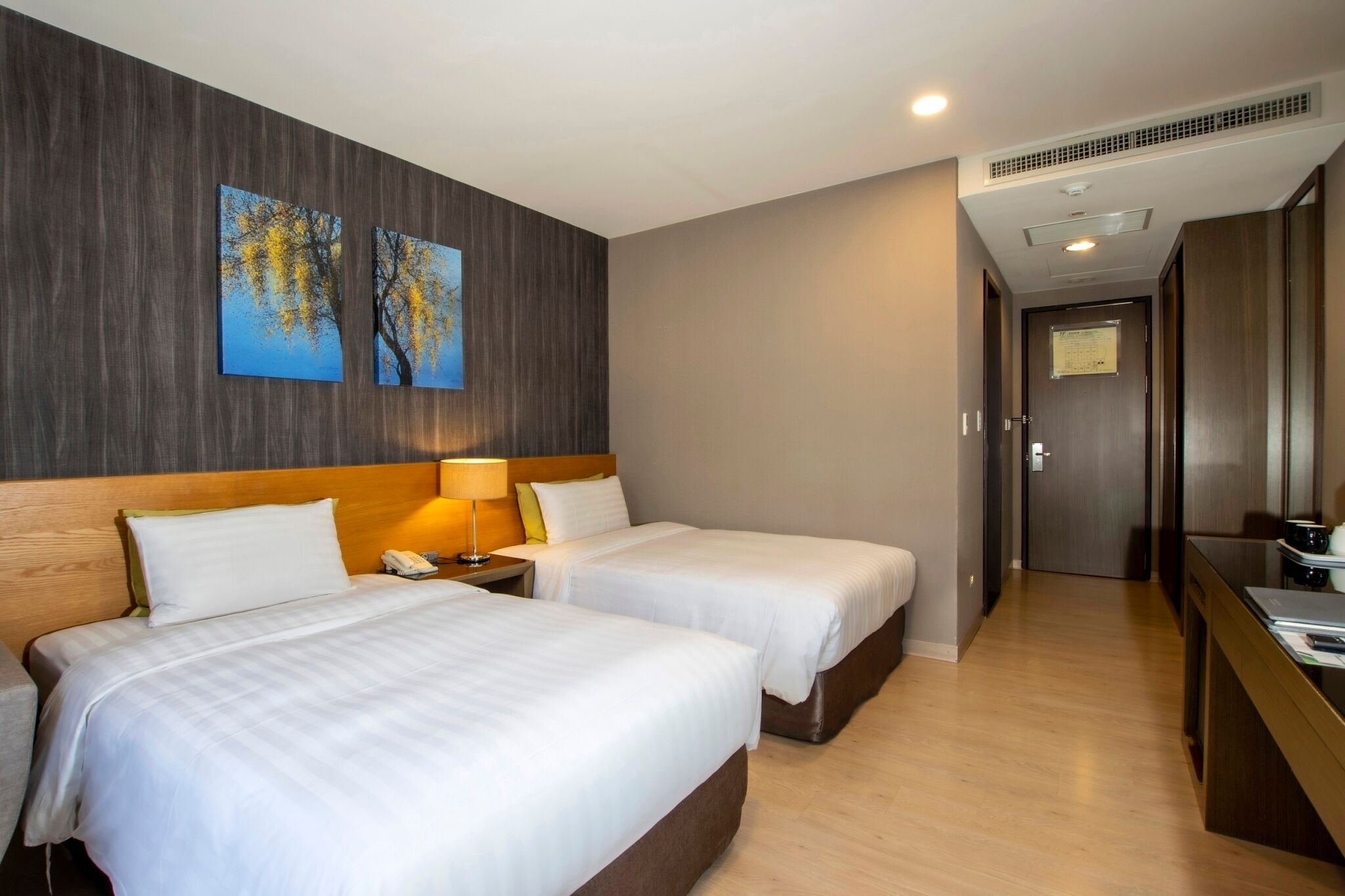 Habitación de hotel con dos camas, cabecera de madera, mesita de noche con lámpara y vista al pasillo.