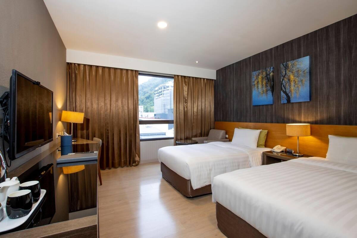 Een hotelkamer met twee bedden, een bureau met televisie, een nachtkastje met lamp en een raam met gordijnen.