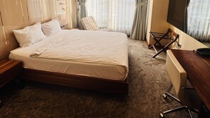 Ein Hotelzimmer mit einem großen Bett, einem Schreibtisch mit Stuhl, einem Fernseher und einem Fenster mit Vorhängen.