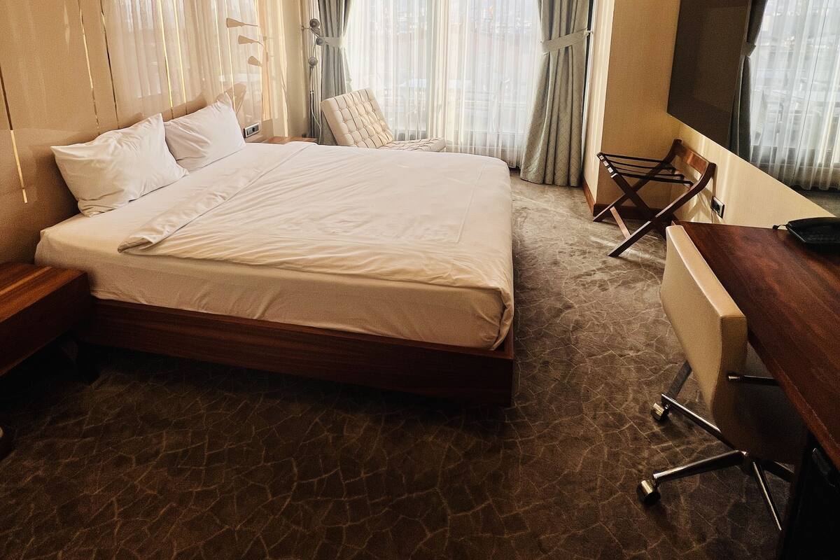 A hotel room with a large bed, a desk with a chair, a TV, and a window with curtains.
