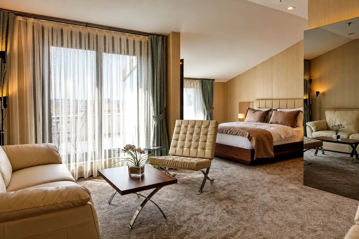 Habitación de hotel con una cama, dos sillones, una mesita de centro y vistas de la ciudad a través de amplias ventanas.