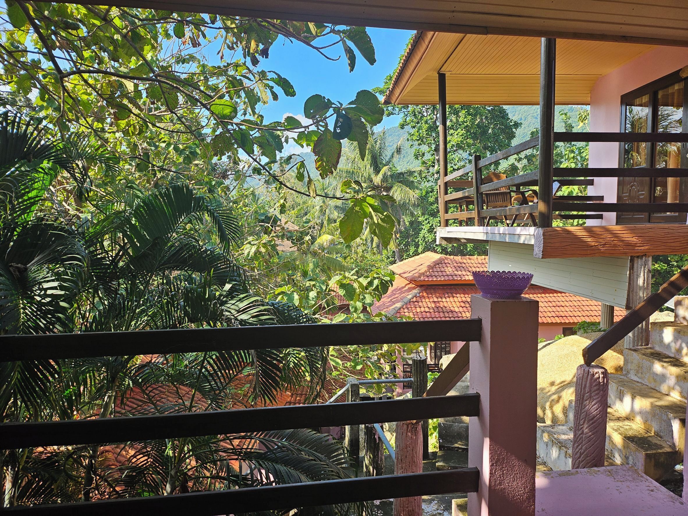 A view from a balcony with a wooden railing, overlooking a lush garden and a building with a tiled roof.