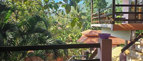 A view from a balcony with a wooden railing, overlooking a lush garden and a building with a tiled roof.