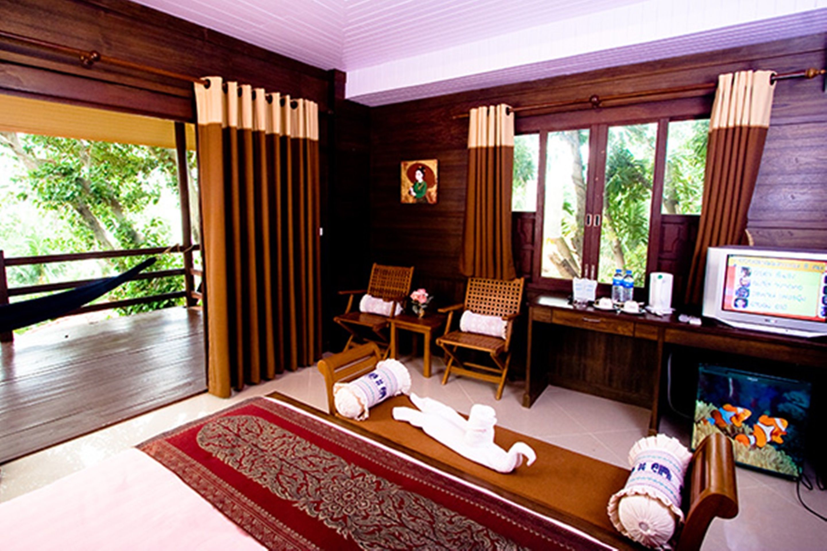 A room with a wooden floor, a red patterned rug, two wooden chairs, a desk with a computer, a television, and an aquarium.