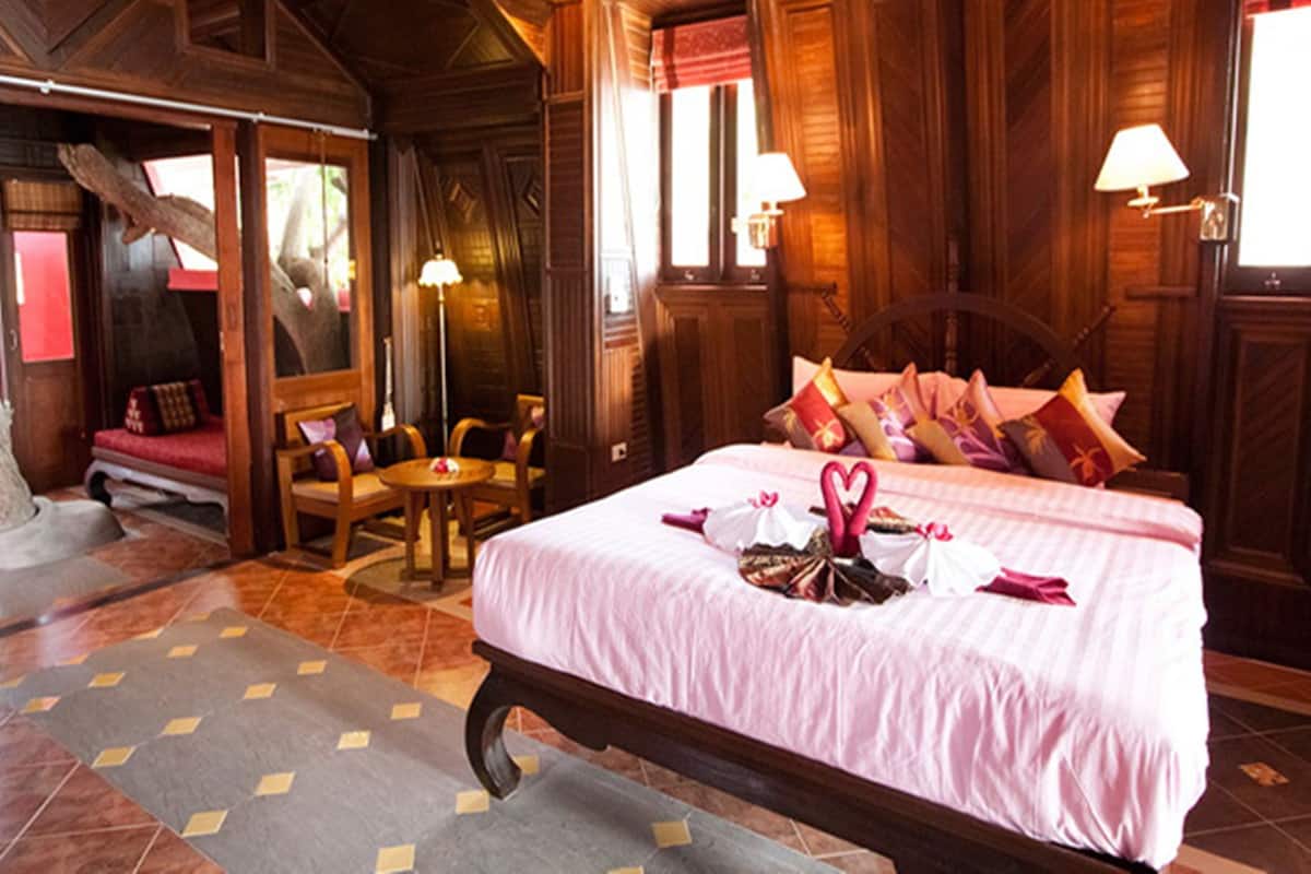A wooden room with a large bed, two chairs, a small table, and a window with curtains.