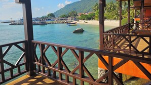 Une terrasse en bois donnant sur une plage, avec des bateaux, une jetée et une chaîne de montagnes en arrière-plan.