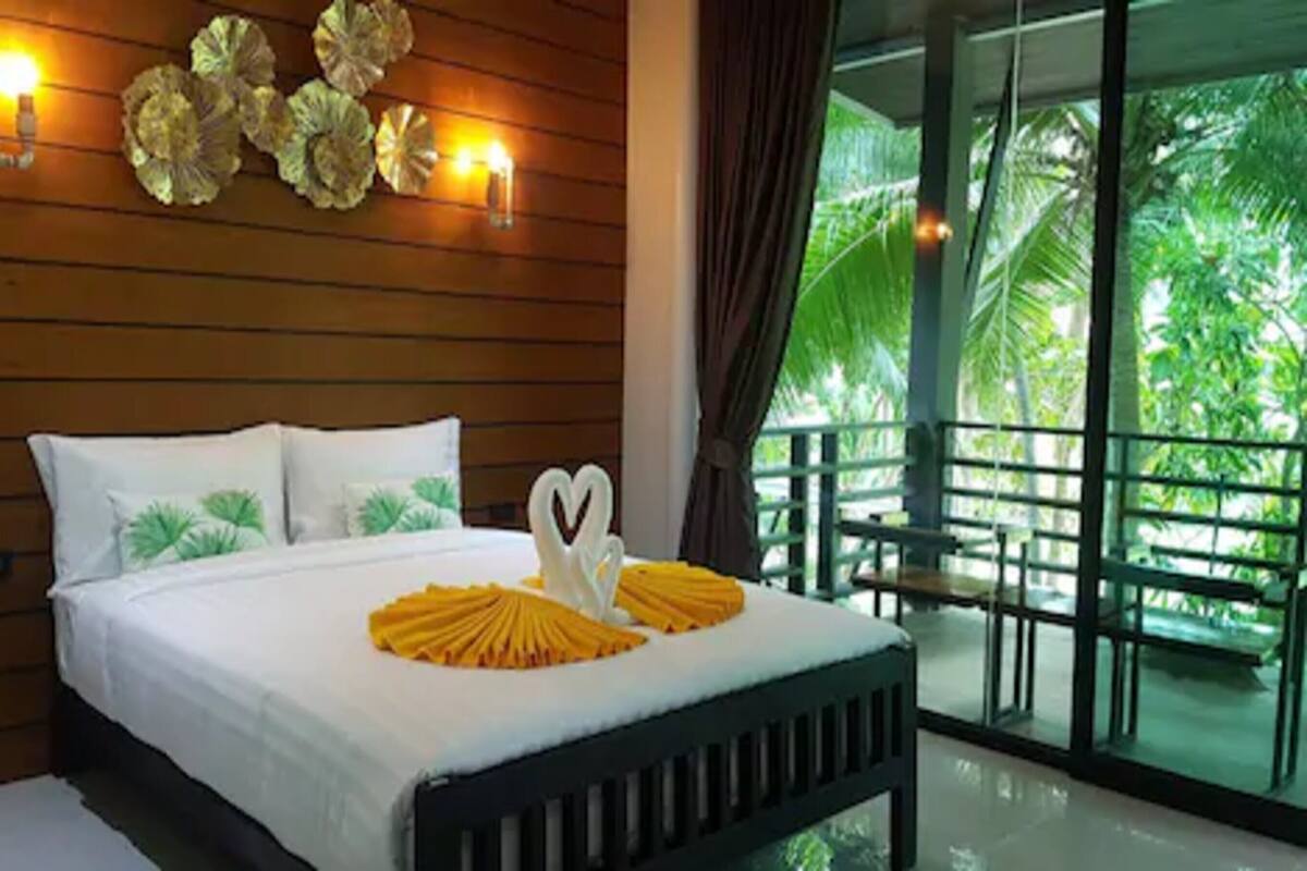 A neatly made bed with white linens and decorative pillows, a heart-shaped centerpiece, and a balcony with a view of lush greenery.