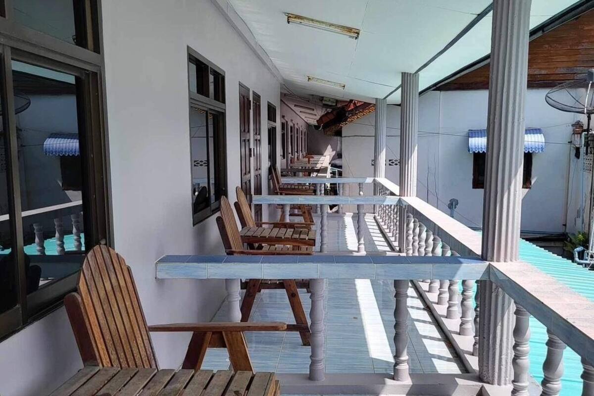 A covered outdoor seating area with wooden benches and a tiled floor, leading to a building with windows and a balcony.
