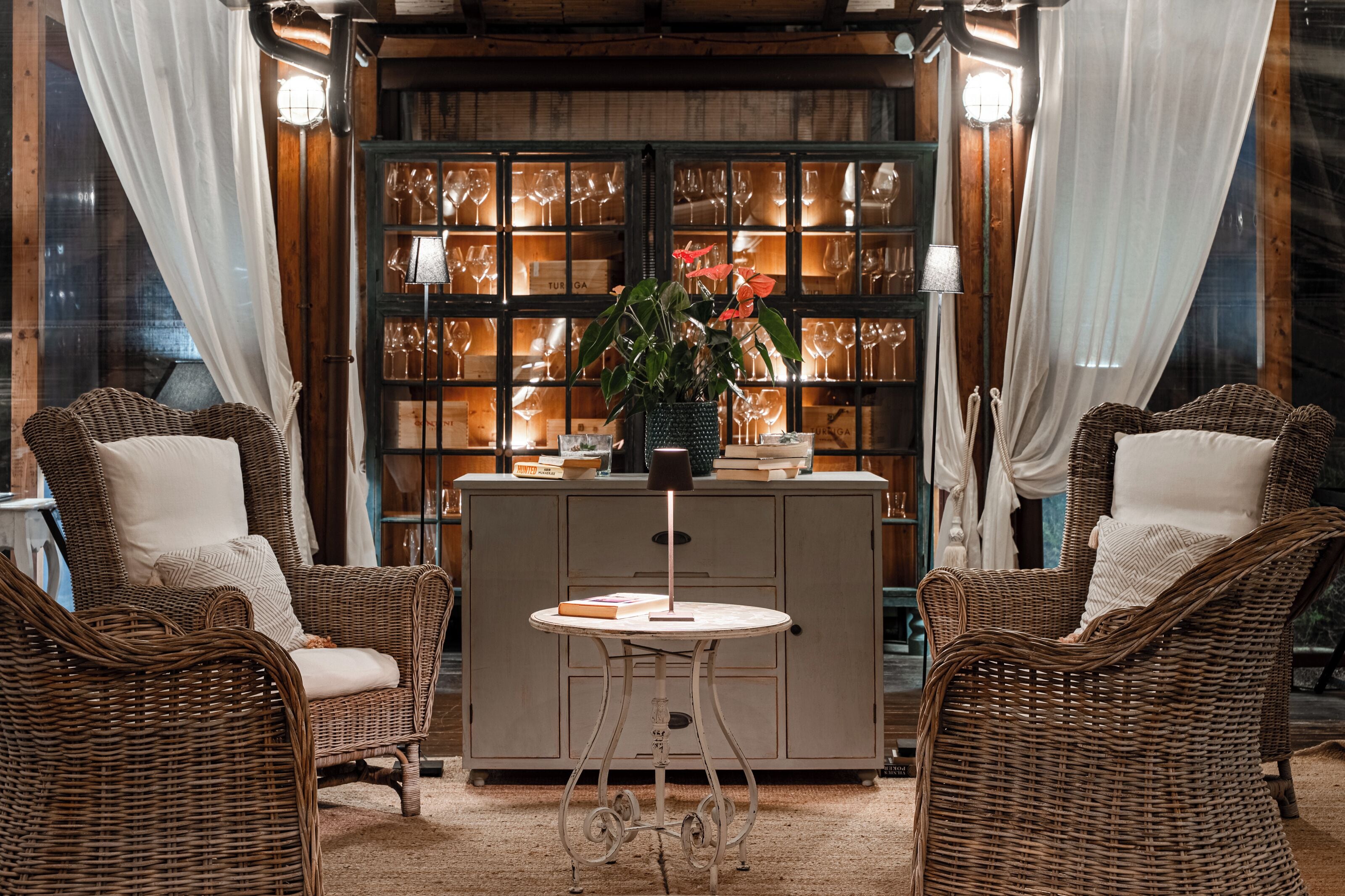 A room with wicker furniture, a glass cabinet with wine glasses, and a table with a lamp.