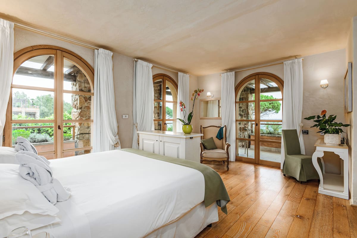 A bedroom with a large bed, two chairs, a small table, and a view through arched windows.