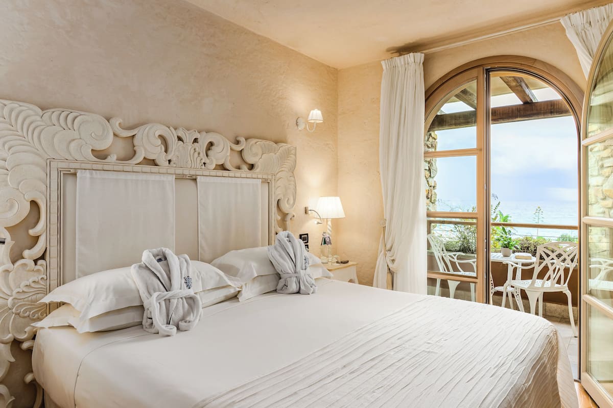 A hotel room with a large bed, two white bathrobes, a nightstand with a lamp, and a balcony with chairs and a table.
