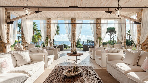 A spacious outdoor seating area with white sofas and a rustic coffee table, set against a backdrop of stone walls and a view of the sea.