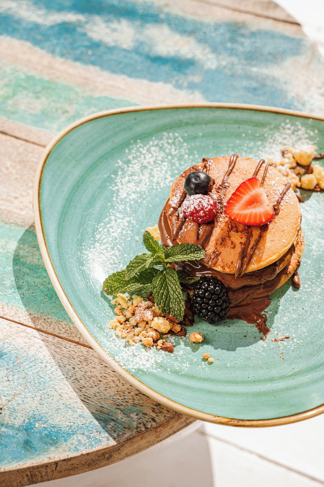 A stack of pancakes with fruit and chocolate sauce on a teal plate.