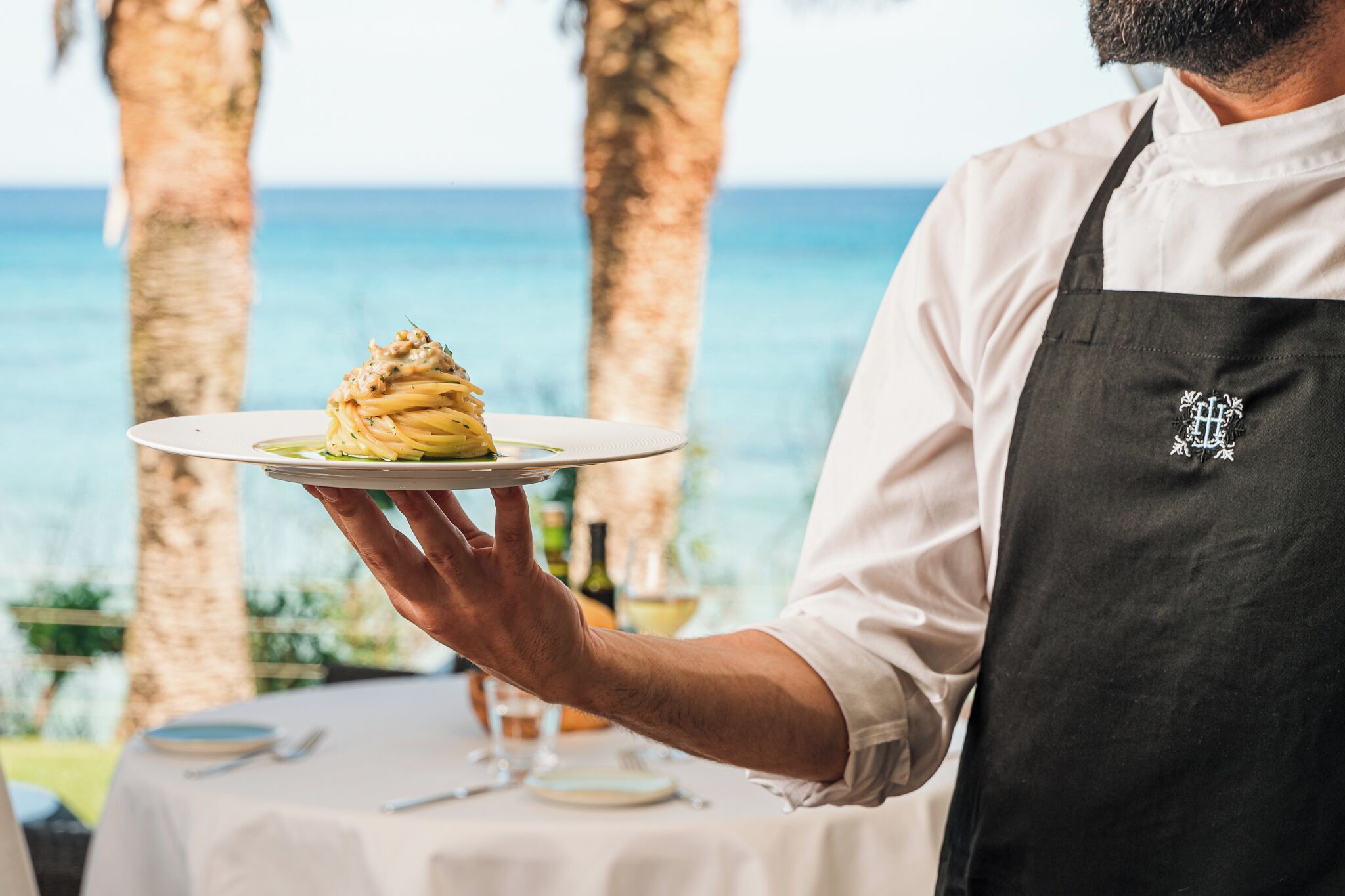 Une personne tenant une assiette de pâtes, portant un tablier de chef, et une table avec une bouteille et un verre en arrière-plan.