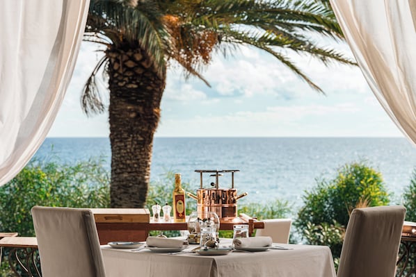 A table set for dining with a view of the sea, a palm tree, and a copper pot.