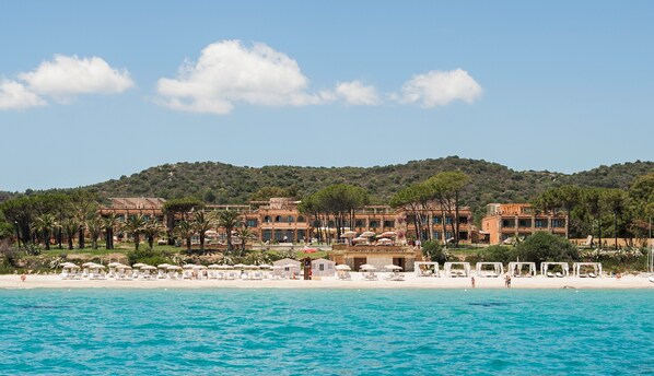Un hotel frente al mar con varias construcciones, una playa y montañas al fondo.