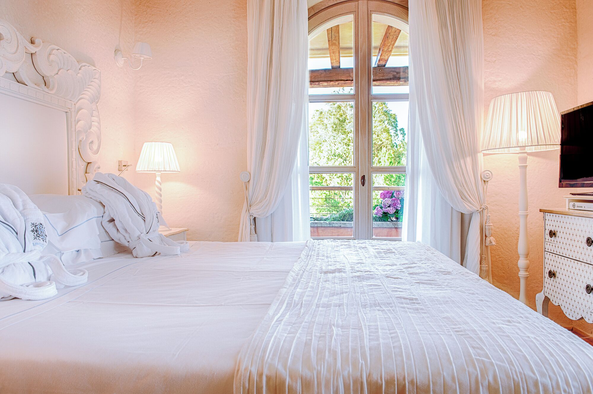 Ein Schlafzimmer mit Bett, Lampe, Fernseher und Blick auf Grün hinter einem Fenster.