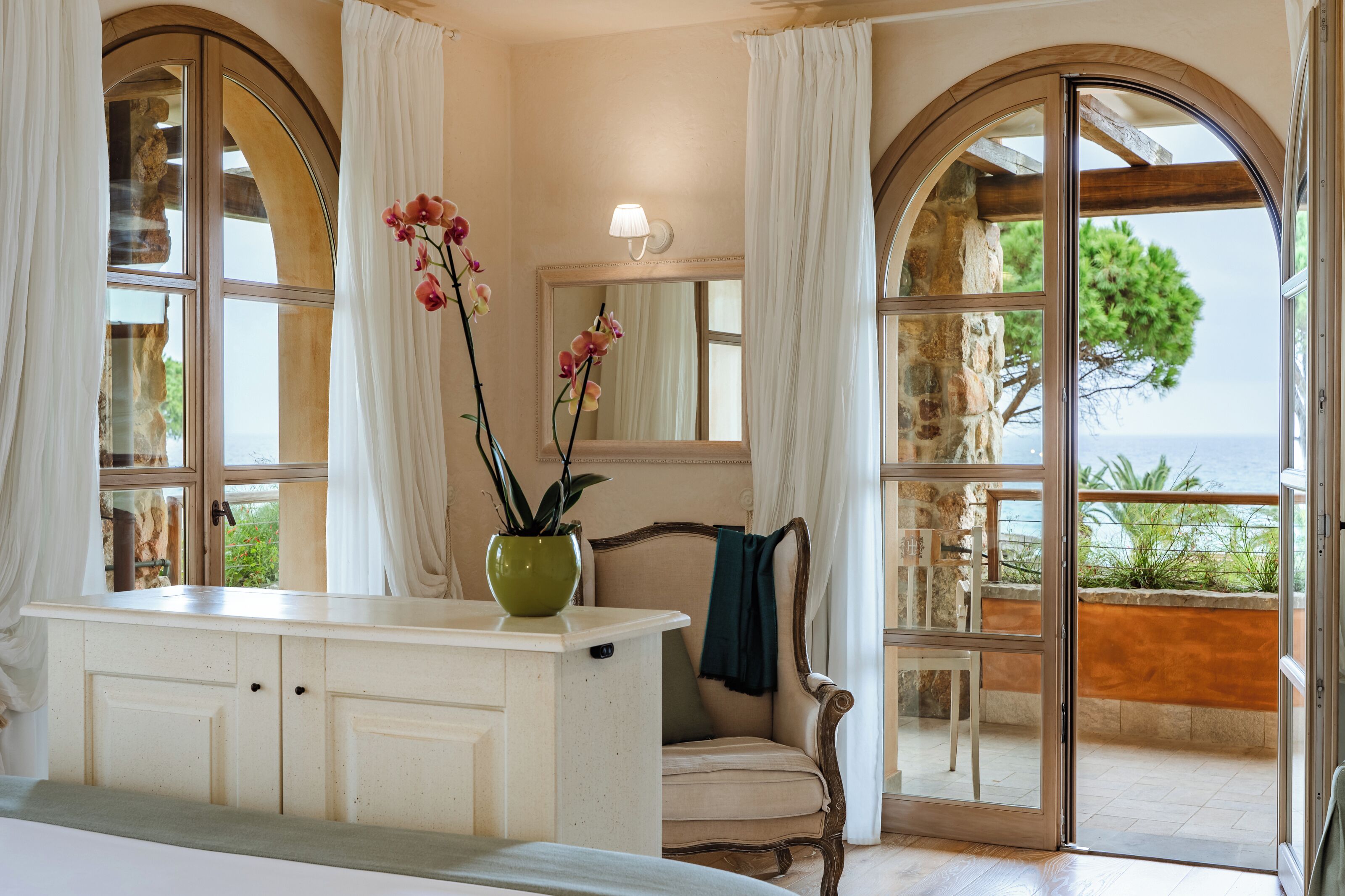 A bathroom with a large arched window, a vanity with a green vase, and a chair by the window.