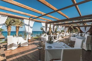 An outdoor dining area with wooden beams, white tablecloths, and chairs, overlooking the sea.