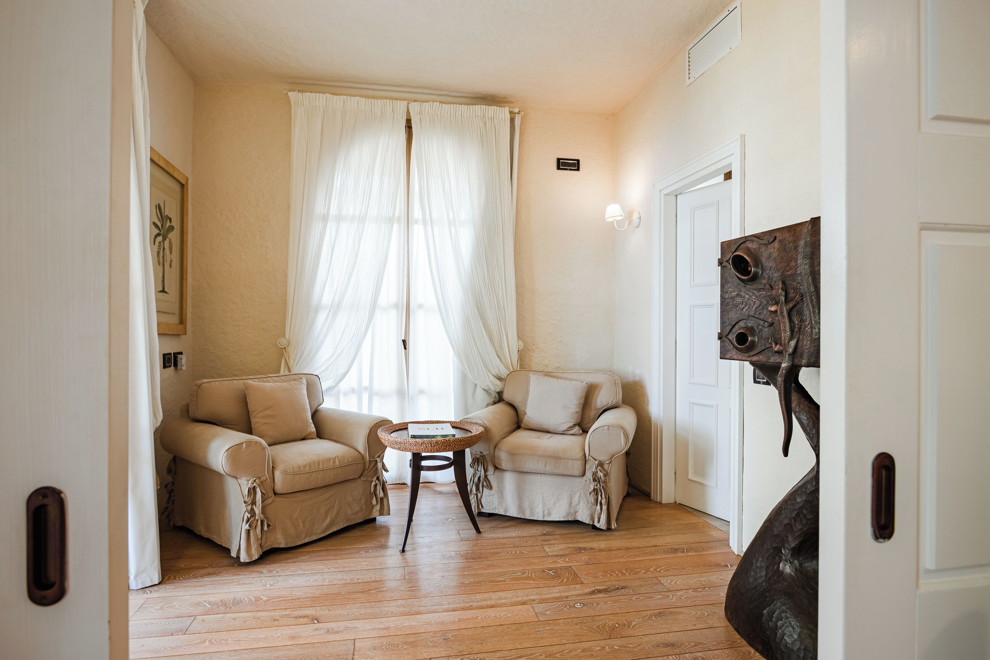 Une pièce comprenant deux fauteuils beiges, une petite table ronde, une grande fenêtre avec des rideaux légers et une porte ornée d’une sculpture en métal.