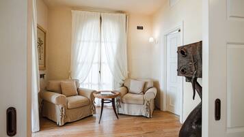 Une pièce comprenant deux fauteuils beiges, une petite table ronde, une grande fenêtre avec des rideaux légers et une porte ornée d’une sculpture en métal.
