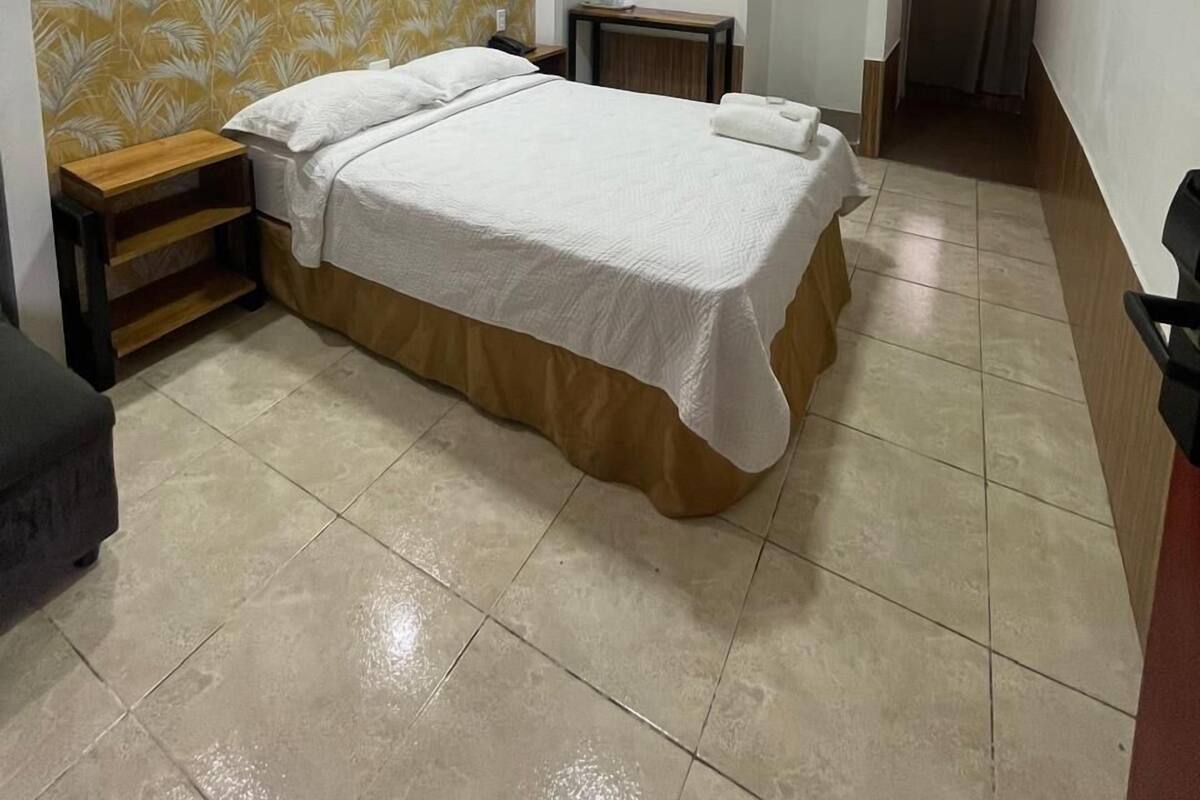 A hotel room with a bed, a nightstand, a mirror, an air conditioning unit, and a television.
