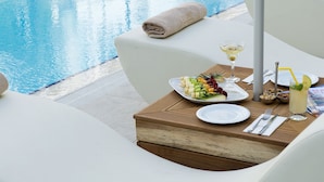 Un área de comedor junto a la piscina con sillas reclinables blancas, una mesa con un plato de comida, una bebida y una botella de agua.