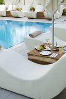 Un área de comedor junto a la piscina con sillas reclinables blancas, una mesa con un plato de comida, una bebida y una botella de agua.