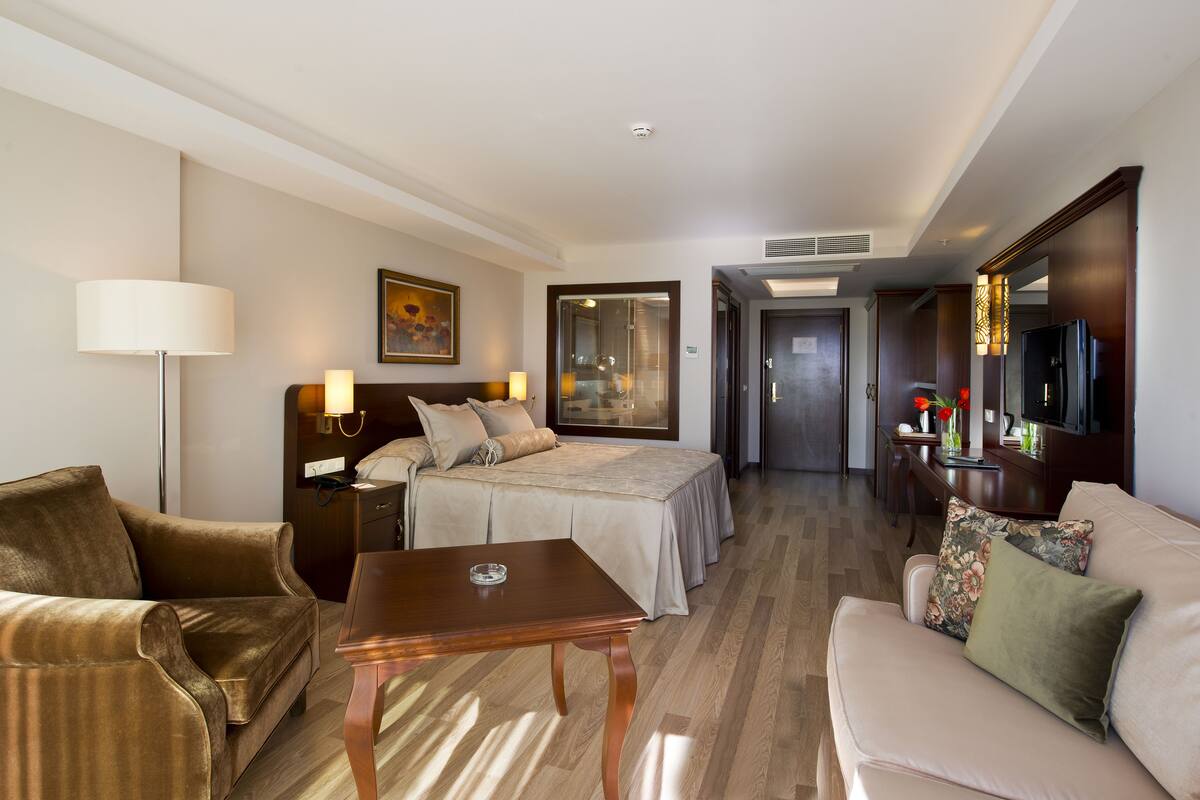 A hotel room with a bed, two sofas, a table, a TV, and a bathroom visible through a glass door.