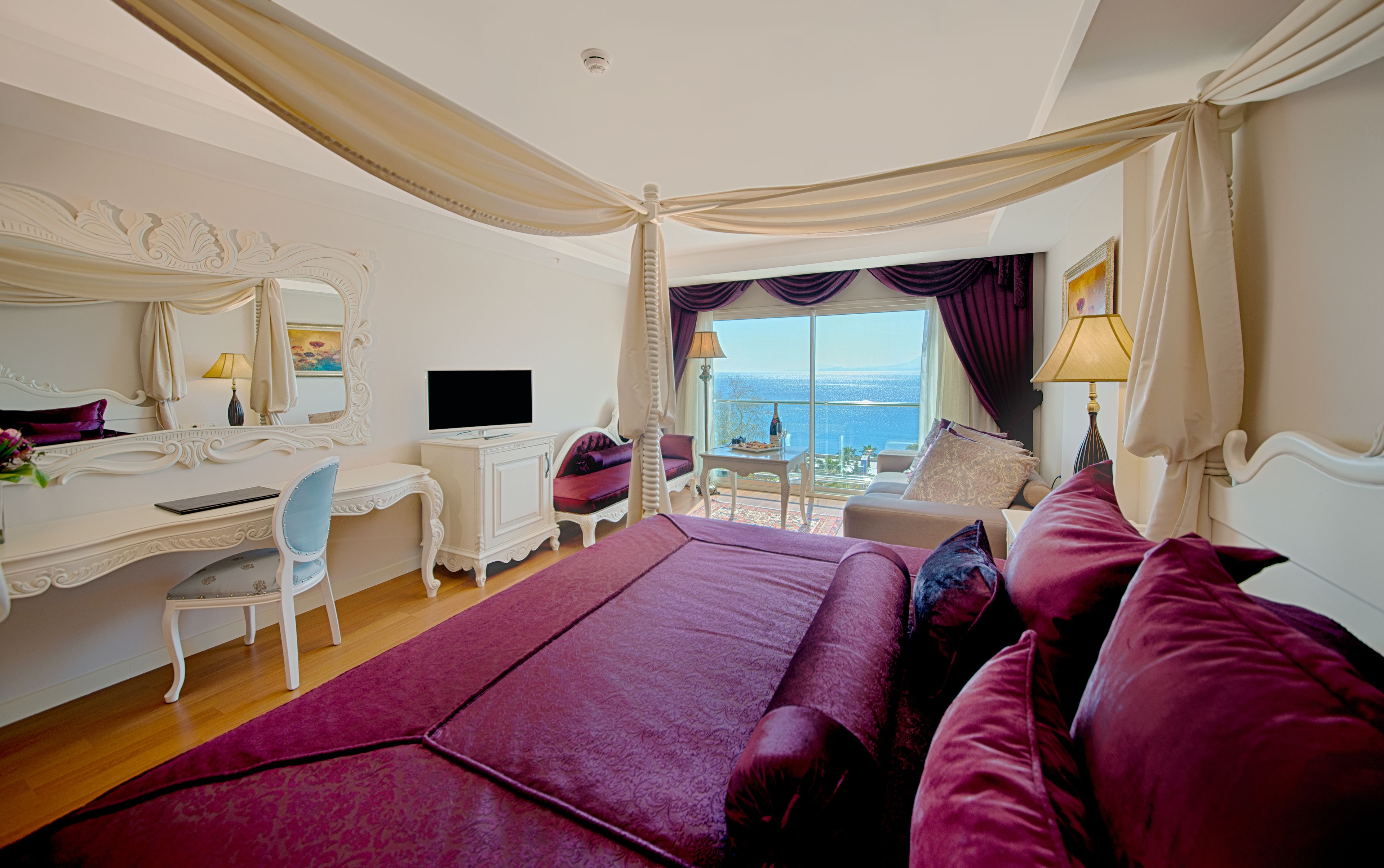 A bedroom with a large bed, a desk, a chair, a TV, and a view of the sea.