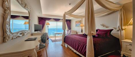 A bedroom with a four-poster bed, a vanity with a mirror, a desk, and a view of the sea.