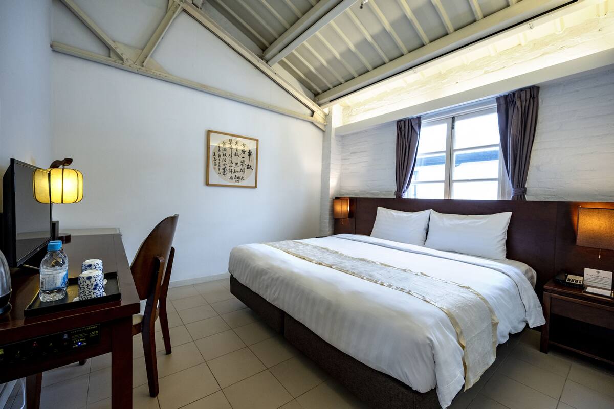 Une chambre d’hôtel comprenant un lit, un bureau avec une lampe, une chaise, une table de chevet et une fenêtre avec des rideaux.