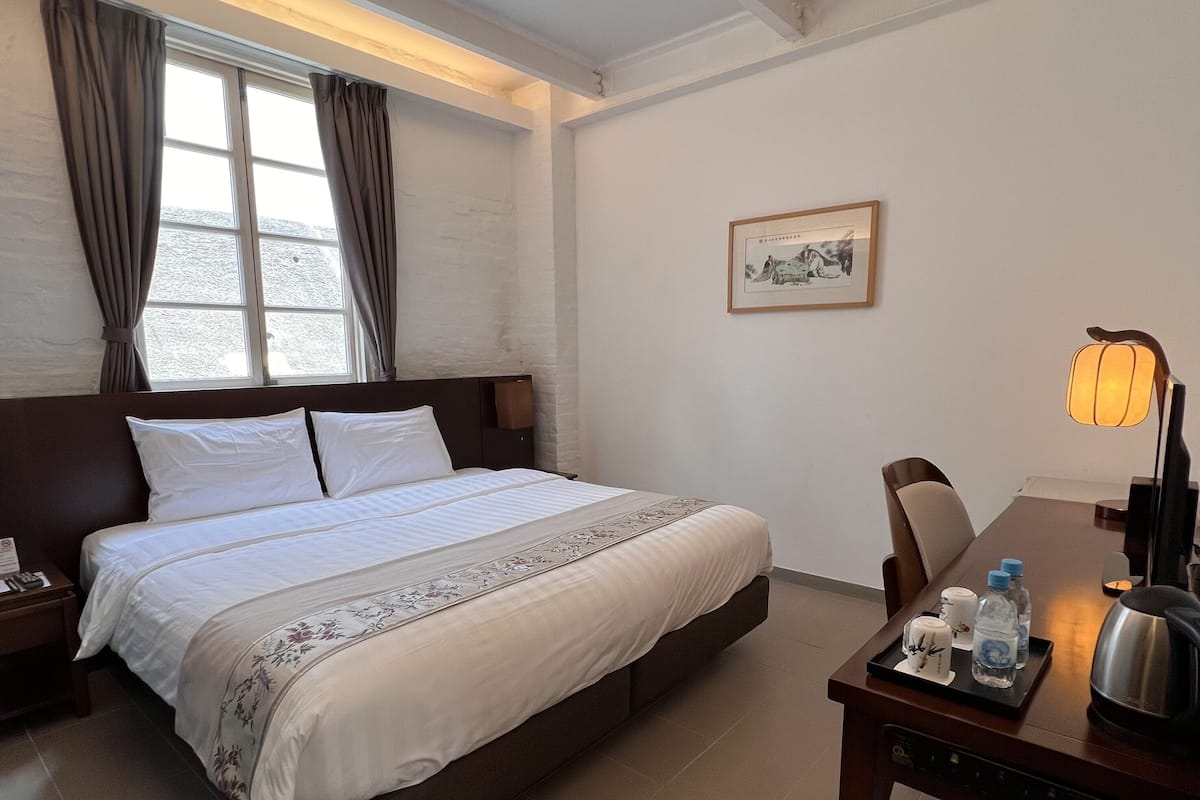Une chambre d’hôtel avec un grand lit, une table de chevet avec une lampe, une chaise et une petite table sur laquelle se trouvent une bouilloire et des bouteilles.