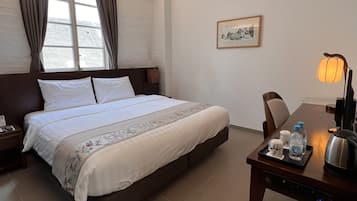 A hotel room with a large bed, a bedside table with a lamp, a chair, and a small table with a kettle and bottles.