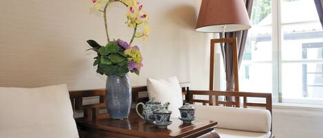 Une table en bois ornée d’un vase de fleurs, d’une théière et de tasses, d’une lampe, et une fenêtre avec des rideaux.