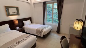 Une chambre d’hôtel avec deux lits, un bureau, une chaise, une lampe, un tableau encadré au mur et une fenêtre avec des rideaux.
