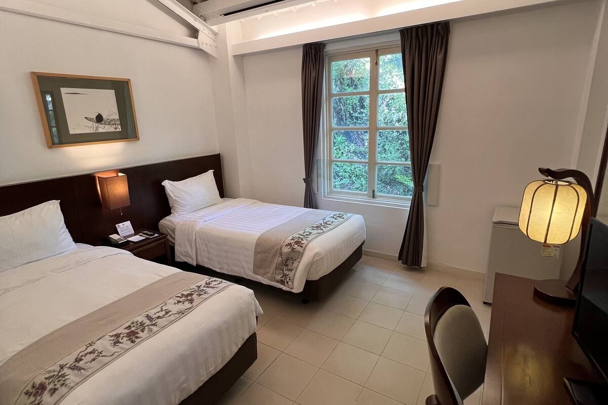 Une chambre d’hôtel avec deux lits, un bureau, une chaise, une lampe, un tableau encadré au mur et une fenêtre avec des rideaux.