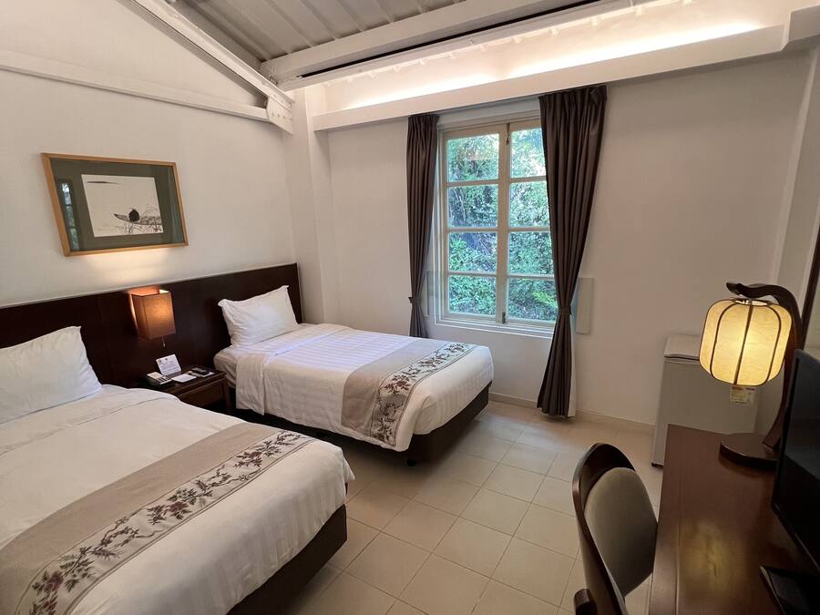 A hotel room with two beds, a desk, a chair, a lamp, a framed picture on the wall, and a window with curtains.