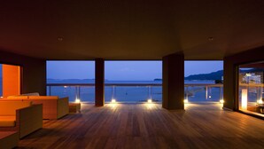 A spacious balcony with wooden flooring, seating area, and large windows offering a view of the sea and mountains.