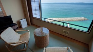 A room with a large window overlooking a calm sea and a rocky breakwater. There are two wicker chairs and a small round table.